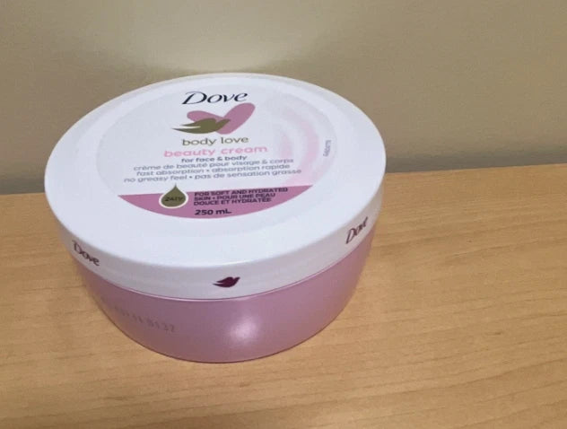 A 250 mL tub of Dove Body Love Beauty Cream for Face & Body, featuring a white lid with pink and green graphics and a pink container base, sitting on a light wood surface against a beige wall.