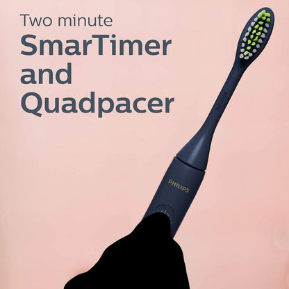 A Philips electric toothbrush with a dark navy handle and green-and-white bristles, shown against a soft pink background with promotional text reading 'Two minute SmarTimer and Quadpacer.' A black hand is holding the toothbrush.
