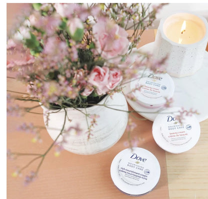 Three round Dove body care jars arranged on a light wood surface beside a white vase with pink flowers and a lit candle. The product labels are visible, including Dove and body cream text.